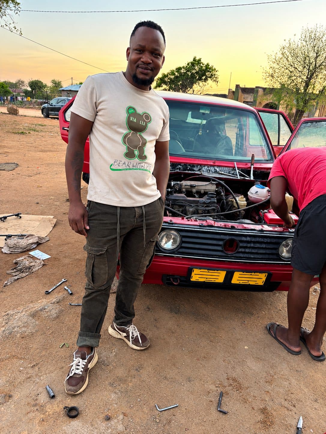 Crecias next to a red VW Golf with its bonnet open, being repaired at sunset